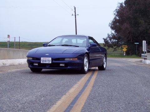 1995 Ford Probe - A blue 1995 Ford Probe is captured from a low angle on a road, with a clear view of its front and side.