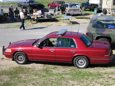 1991 Ford LTD Crown Victoria - A red police sedan with a light bar is parked on a dirt and grass area, with other vehicles in the background.