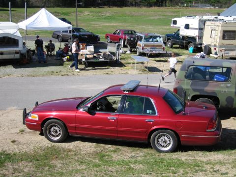 1991 Ford LTD Crown Victoria - A red police car with a light bar is parked on the side of the road, with other vehicles and tents in the background.