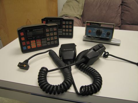 1991 Ford LTD Crown Victoria - Two GE radio control heads beside a Federal Signal siren controller, with two black handheld mics and tangled coiled cords on a table.