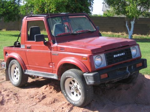 1987 Suzuki Samurai - This red 1987 Suzuki Samurai is shown from a three-quarter front angle, with the 'SUZUKI' text visible on the grille.