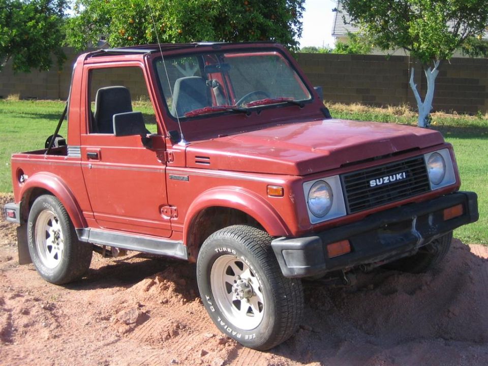 Red 1987 Suzuki Samurai with a soft top, parked on dirt. The grille has 'SUZUKI' text