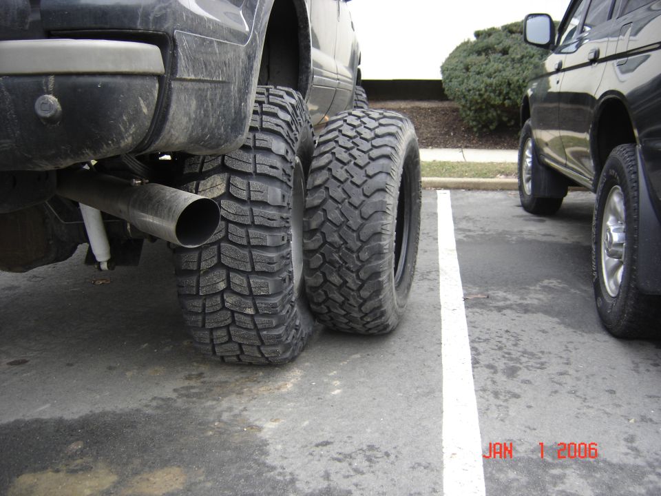Rear view of a dark SUV with large off-road tires and a prominent exhaust pipe, parked next to another vehicle