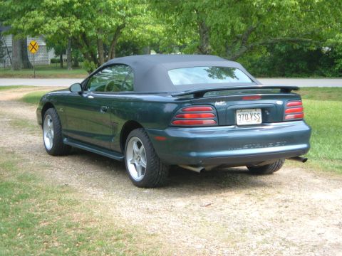 1995 Ford Mustang - A green convertible Mustang is parked on a gravel driveway with trees in the background.