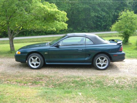 1995 Ford Mustang - A side view of a dark green convertible Mustang parked outdoors on a gravel surface.