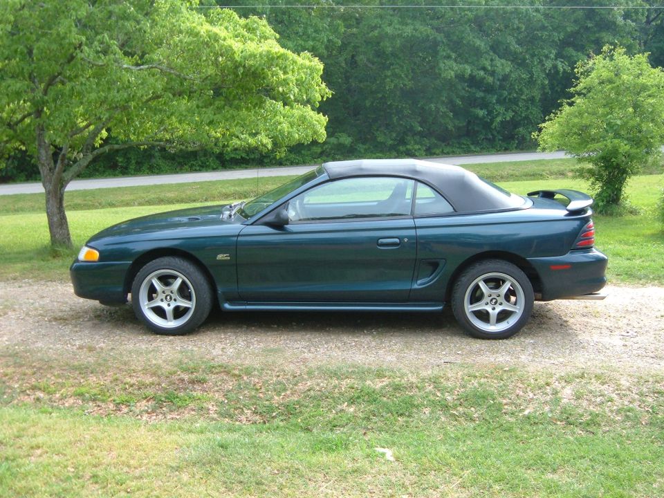 Dark green convertible Mustang parked on a gravel driveway with trees in the background