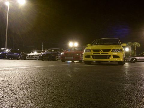 1994 Ford Probe - Several cars are parked in a lot at night, with a yellow Mitsubishi Lancer Evolution in the foreground.