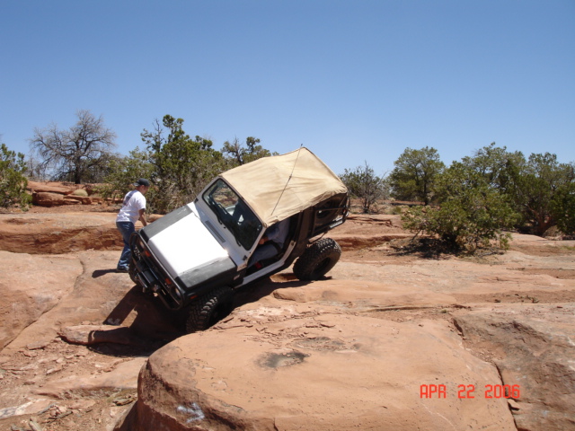 White Jeep with tan soft top tipped on slickrock as a man spots from the side on red rock trail