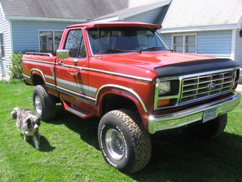 1984 Ford F-150 - A red 1984 Ford F-150 pickup truck is parked on a grassy lawn next to a house, with a dog standing nearby.
