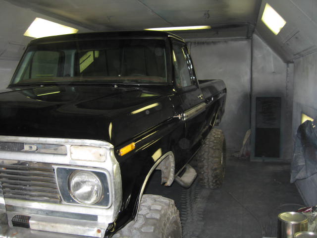 Black classic Ford pickup with chrome grille parked inside a gray spray booth, lifted on mud tires