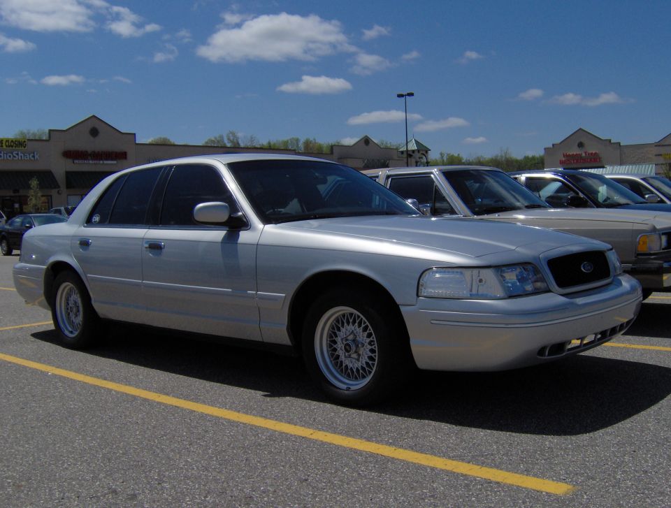 Silver 2002 Ford Crown Victoria parked in a lot, front passenger side visible. Other cars are parked nearby