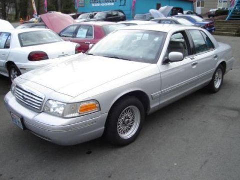 2002 Ford Crown Victoria - A silver 2002 Ford Crown Victoria is parked in a lot with other vehicles and a building in the background.
