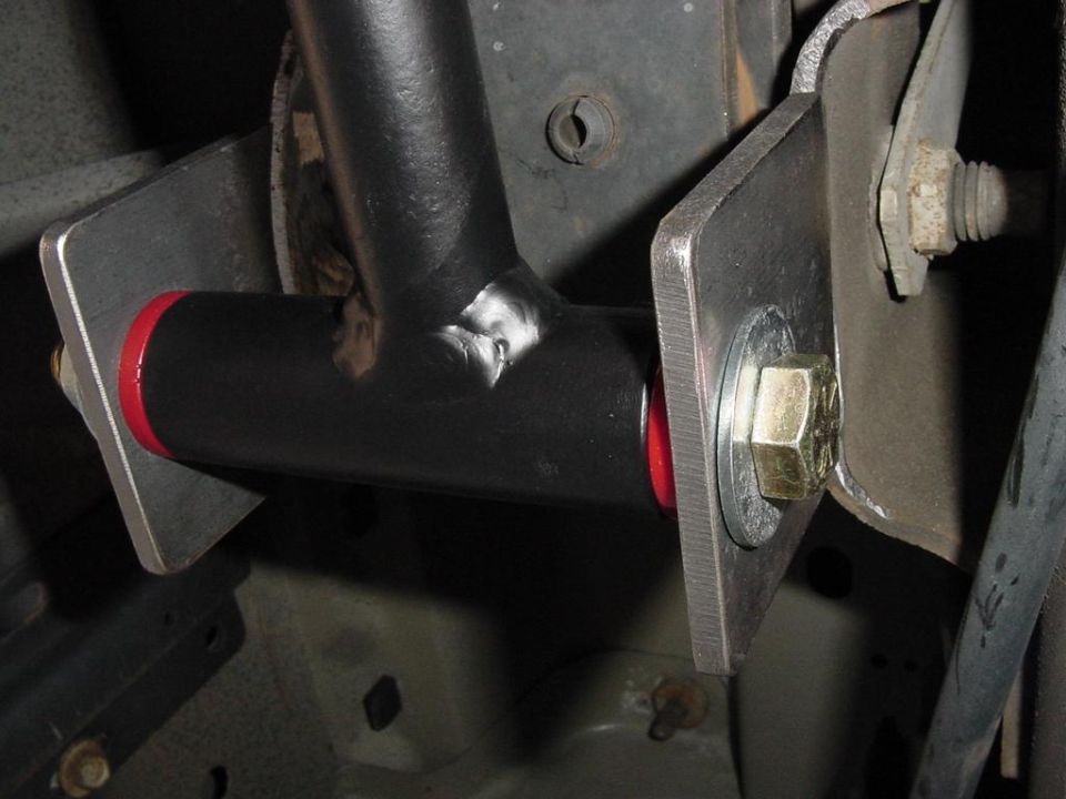 Black suspension link with red poly bushings bolted between steel brackets under a vehicle