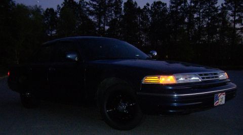 1996 Ford Crown Victoria - A dark sedan with black wheels is parked outdoors at dusk, with trees in the background.