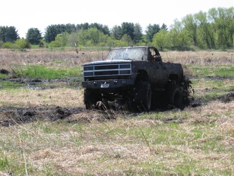 1977 Ford F-150 - A heavily mud-splattered pickup truck navigates a challenging off-road course, kicking up dirt and mud.