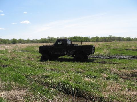 1977 Ford F-150 - A pickup truck is covered in mud while stuck in a field with a blue sky and trees in the background.