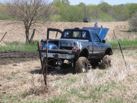 1977 Ford F-150 - A blue pickup truck is stuck in a muddy field with its hood open, while another vehicle is visible in the distance.