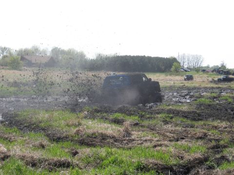 1977 Ford F-150 - A blue SUV is stuck in a mud pit, with mud spraying high into the air. Other vehicles are parked in the background.