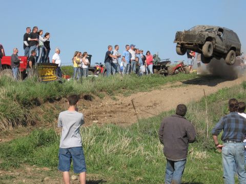 1977 Ford F-150 - A dark-colored SUV is airborne over a dirt ramp, with a crowd of people watching the event.