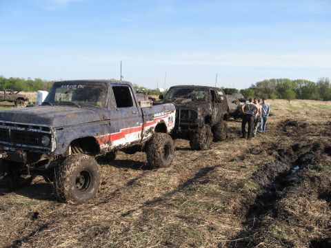 1977 Ford F-150 - A lineup of mud-caked trucks and SUVs are stuck in a field during a mud bogging event.