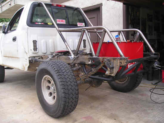 White 2002 Ford F-150 pickup truck chassis with a roll cage and red fuel tank, viewed from the rear quarter