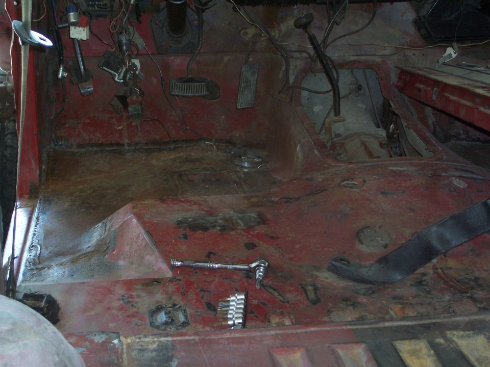 Interior floor of a vehicle with red paint, rust, pedals, and a shifter. Tools are on the floor