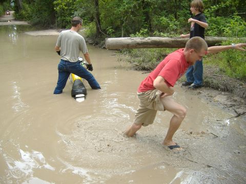 2004 Jeep Wrangler - A motorcycle is stuck in a muddy water crossing, with two people attempting to free it and another person observing.