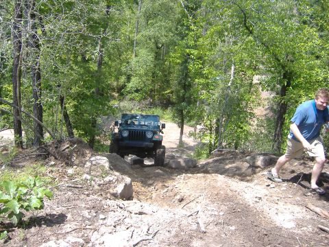 2004 Jeep Wrangler - A green 2004 Jeep Wrangler tackles a challenging, rocky off-road path surrounded by dense trees, with a man ascending the embankment to the…