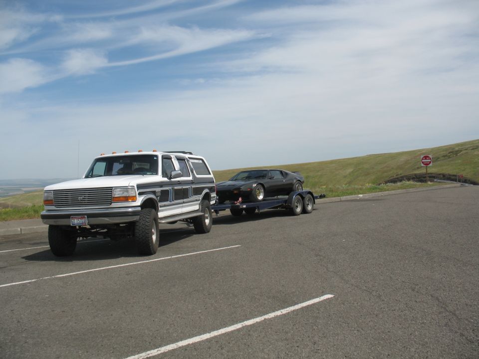 White Ford truck towing black DeTomaso Pantera on a flatbed trailer in a hilltop parking lot.
