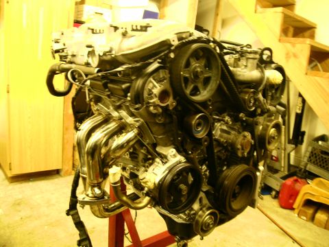 1995 Ford Probe - V6 engine on a red engine stand in a garage, viewed from the accessory-drive end with the timing belt, cam pulleys, crank pulley and power-…
