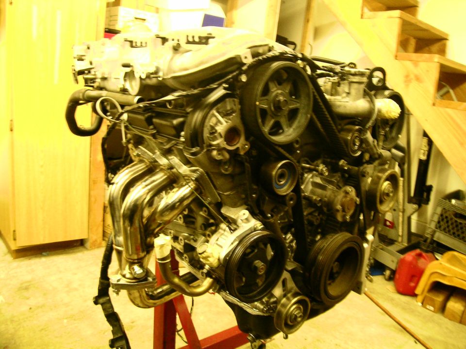 V6 engine on red stand with stainless tubular header and exposed timing belt in garage