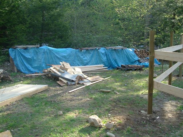 Construction site with lumber, pallets, and blue tarp covering woodpile