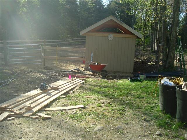 Outdoor construction area with a shed, lumber pile, wheelbarrow, and tools