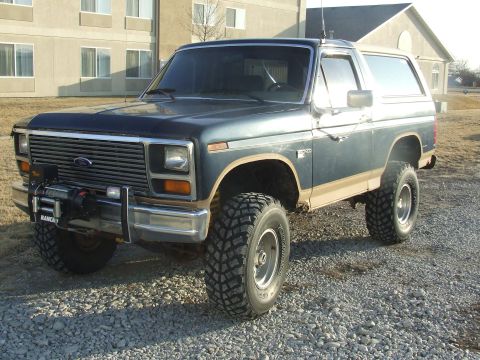 1986 Ford Bronco - Driver-side 3/4 front view of a lifted two-tone dark blue and tan full-size Ford Bronco on a gravel lot, fitted with a Rancho-branded winch…