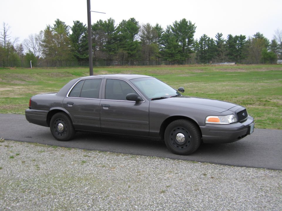 Gray Ford Crown Victoria parked on a paved surface with a grassy field and trees in the background