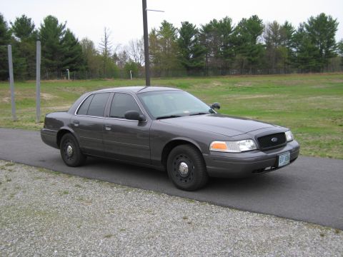1991 Ford LTD Crown Victoria - A dark gray Ford Crown Victoria sedan is parked on a paved surface with a grassy area and trees in the background.