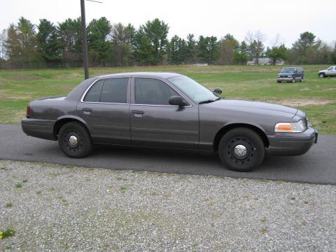 1991 Ford LTD Crown Victoria - A gray Ford Crown Victoria police car is parked on a gravel surface with a field and trees behind it.