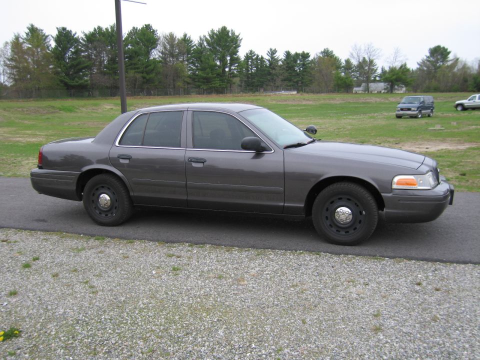 Gray Ford Crown Victoria police car parked on gravel, trees and field in background
