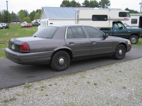 1991 Ford LTD Crown Victoria - A dark gray sedan is parked on a paved surface next to a gravel area, with buildings and other vehicles in the background.