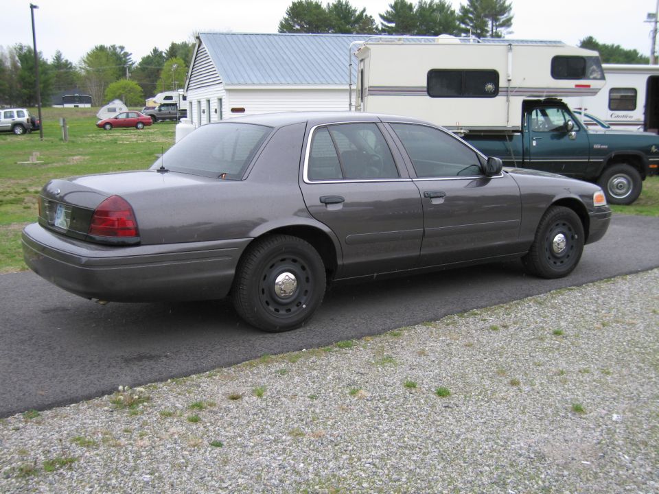 Gray Ford Crown Victoria parked on asphalt with a gravel shoulder. A camper and a green truck are behind it