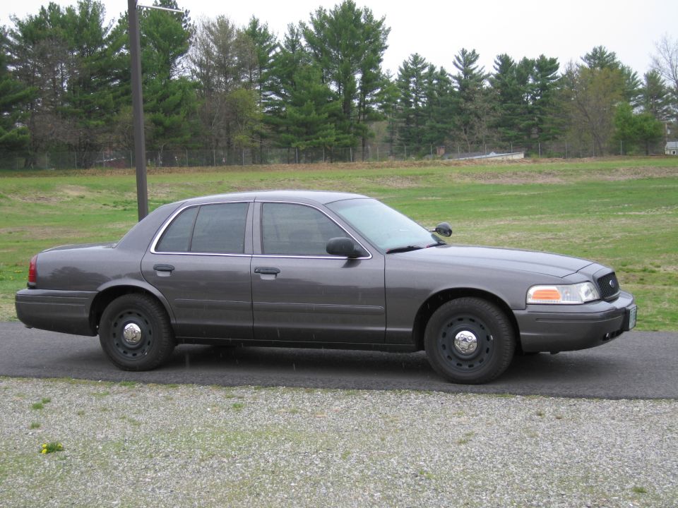 Gray Ford Crown Victoria sedan, side view, parked on asphalt with gravel shoulder
