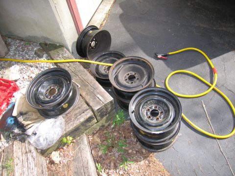 1991 Ford LTD Crown Victoria - A collection of steel wheels are piled on wood, with a yellow garden hose nearby.