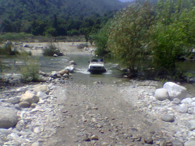 White SUV crossing a rocky riverbed with trees and hills in the background