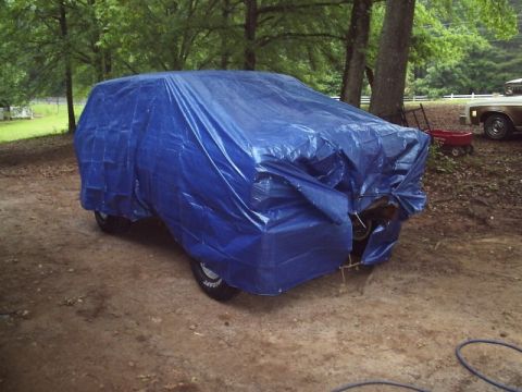 1978 Ford Bronco - A full-size SUV sits on red dirt under trees draped in blue poly tarps tied down with rope, with only the mud-terrain tires and chrome whee…