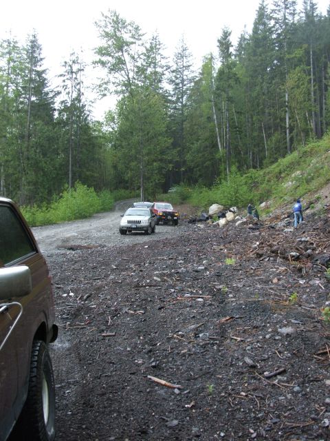 1980 Ford Bronco - Multiple SUVs are parked on a gravel road surrounded by dense forest and trees.