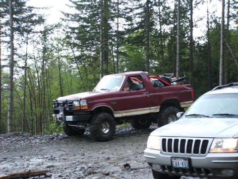 1980 Ford Bronco - A maroon Ford Bronco with off-road modifications is parked on a dirt road, with another vehicle in the foreground.