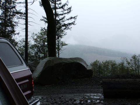1980 Ford Bronco - The rear of a red SUV is visible on a wet, muddy surface, with a backdrop of trees and fog-covered hills.
