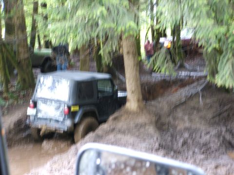 1980 Ford Bronco - Jeep Wrangler navigating a muddy trail in a forest setting with another vehicle nearby.