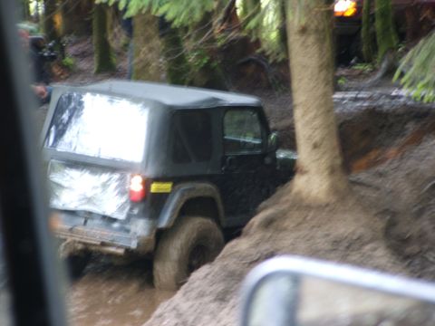 1980 Ford Bronco - A black Jeep Wrangler struggles through a muddy off-road trail surrounded by trees, with another vehicle visible in the background.