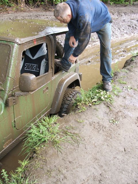 1980 Ford Bronco - A person steps into a mud-covered Jeep Wrangler that is stuck in a muddy ditch.